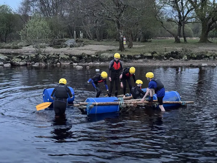 Scouts in yellow helmets and wetsuits build a raft from wooden poles and blue barrels in shallow water.