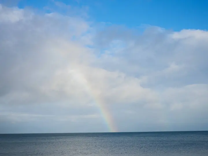 A rainbow over a calm sea under a blue sky and scattered clouds.